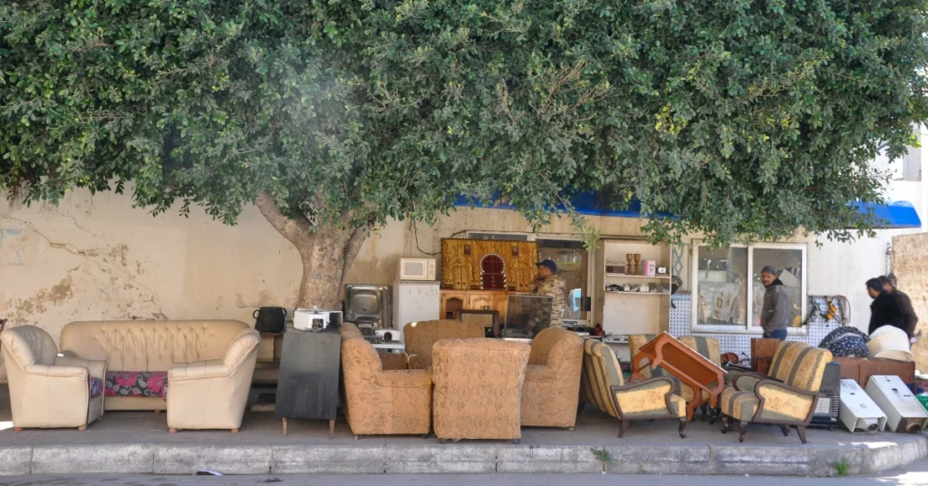 A street-side second-hand shop displays various vintage sofas, armchairs, and household items on the sidewalk under the shade of a large green tree. 📸
